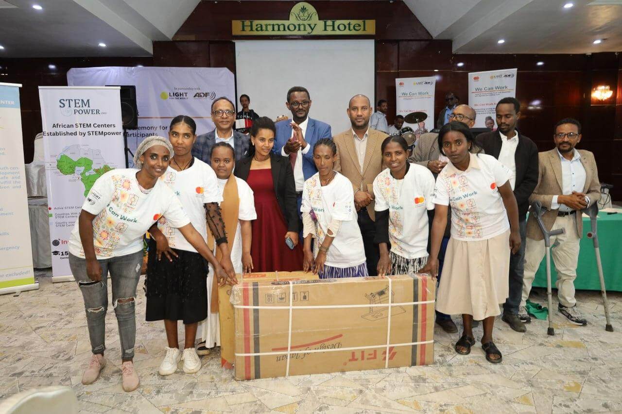 A group of We Can Work graduates and programme staff pose together at the graduation ceremony at Harmony Hotel. Six women in the foreground wear white "We Can Work" T-shirts and stand around a boxed sewing machine. Behind them, several officials and partners smile for the camera. Banners for STEMPower, Light for the World, ADF and the Mastercard Foundation are visible in the background.