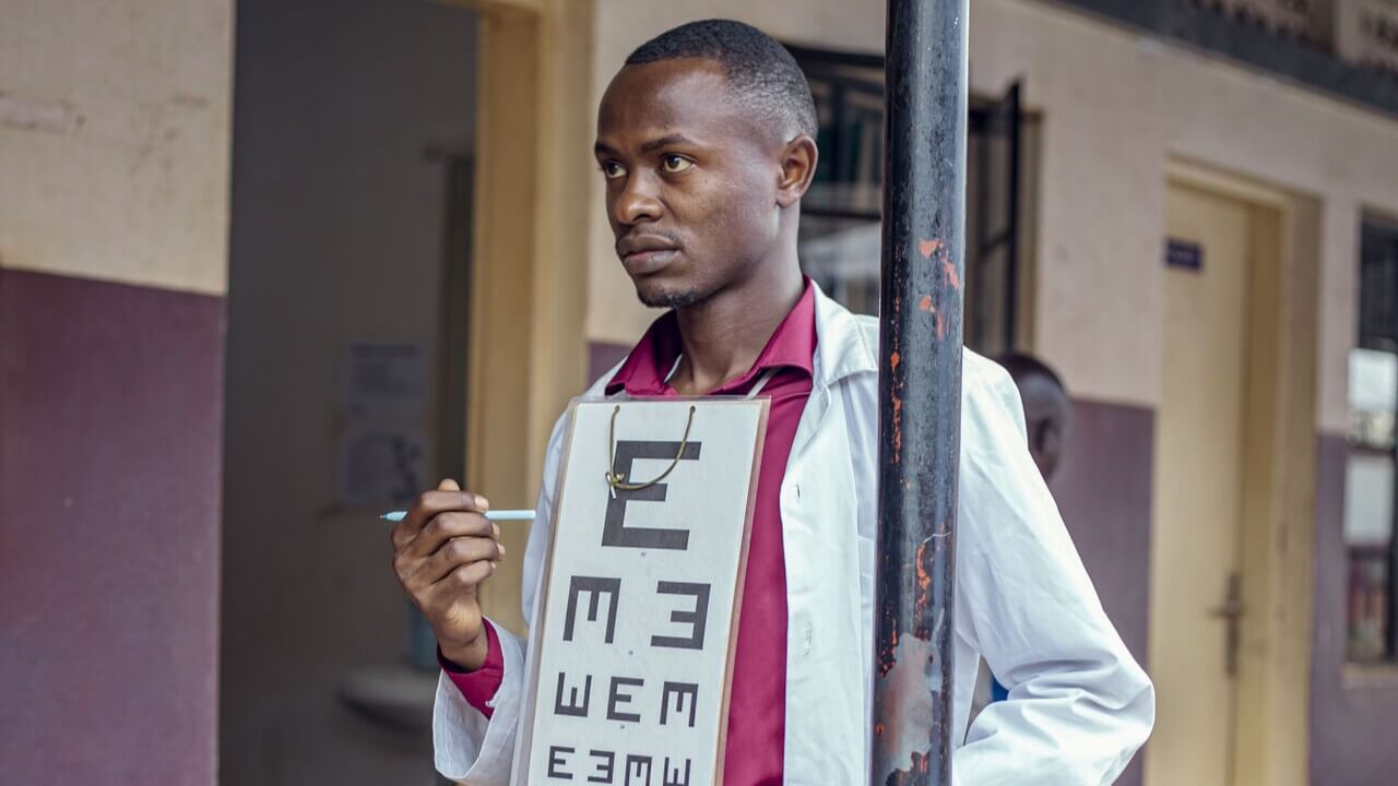 A health worker in a white coat holds a tumbling-E eye chart and a pointer, standing outside a hospital ward.