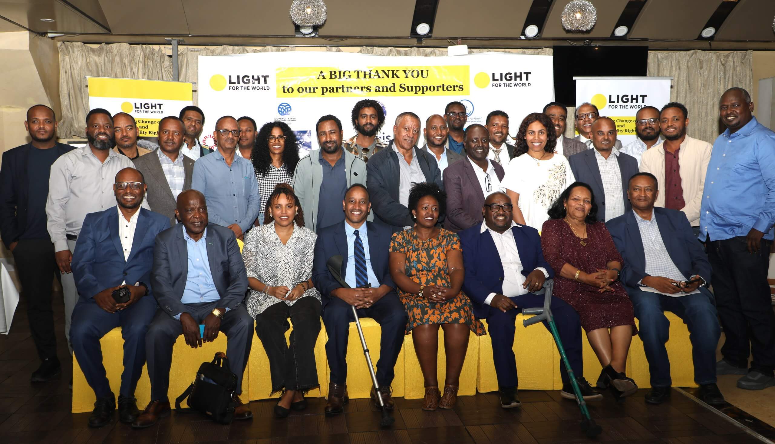 A group of around 30 partners, government representatives and Light for the World staff pose together at the Ethiopia strategy launch. The front row is seated on a yellow bench, with two people holding mobility crutches. Behind them, a banner reads "A big thank you to our partners and Supporters" alongside Light for the World branding.