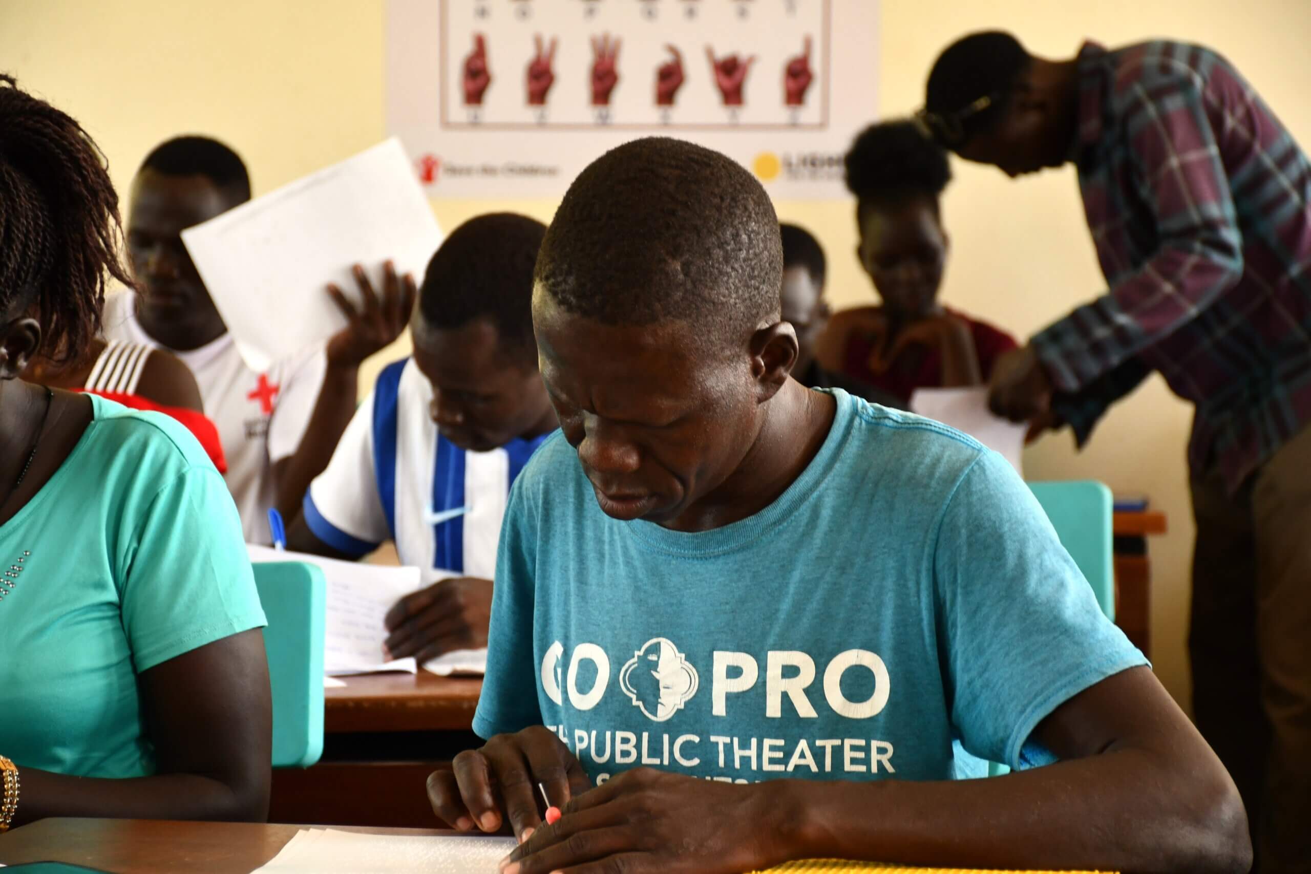 A student teacher in a teal t-shirt writes on a Braille slate at his desk, concentrating as he uses a stylus. Other student teachers sit at nearby desks working on examination papers. A sign language alphabet chart is visible on the wall behind them.