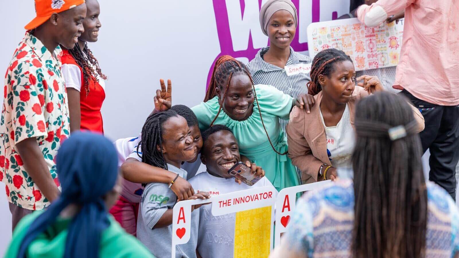 A group of young jobseekers smile, laugh and pose for a phone photo at the Talent Without Limit career fair in Kampala. Some hold large playing-card props from the #PlayTheFullDeck campaign, including an "Ace of Hearts: The Innovator" card. A banner at the front displays Brighter Monday Uganda, the National Council for Persons with Disabilities and Light for the World logos.
