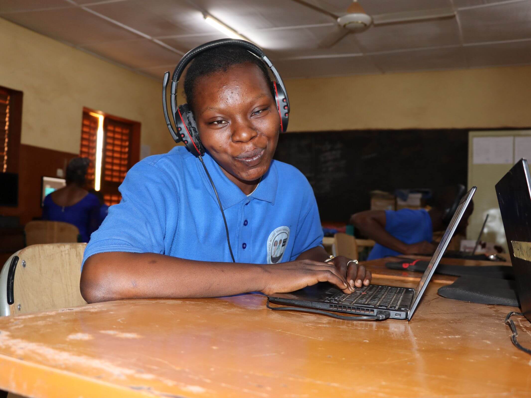 Jannet Aude Compaore wears headphones and types on a laptop in a classroom, smiling at the camera. Other students work at laptops in the background. They are in a classroom in Ouagadougou.