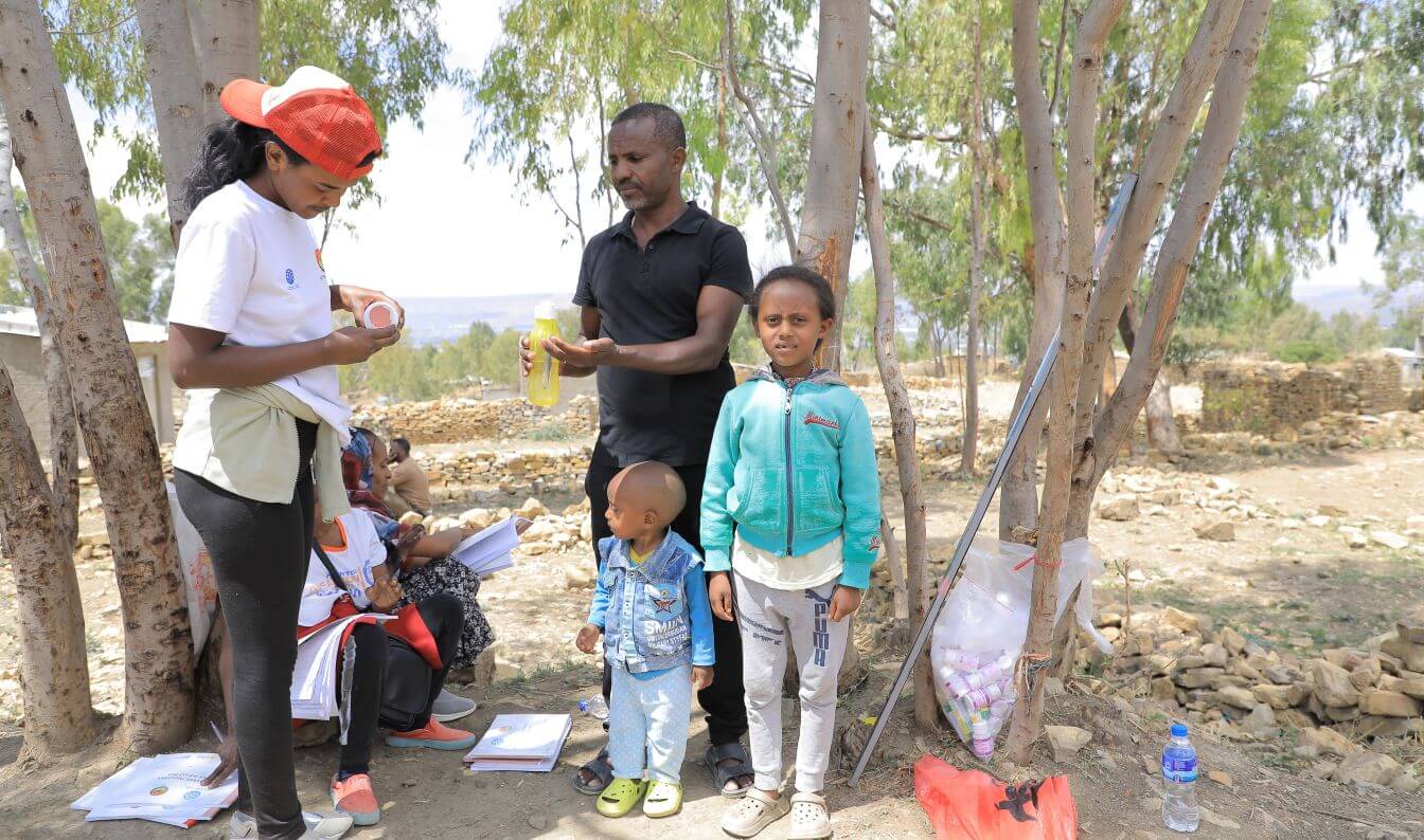 A health worker prepares medication beside Berhane Teklay and his two children, Lidiya Berhanu and her younger brother Temesgen Berhane, outdoors during a trachoma treatment campaign.