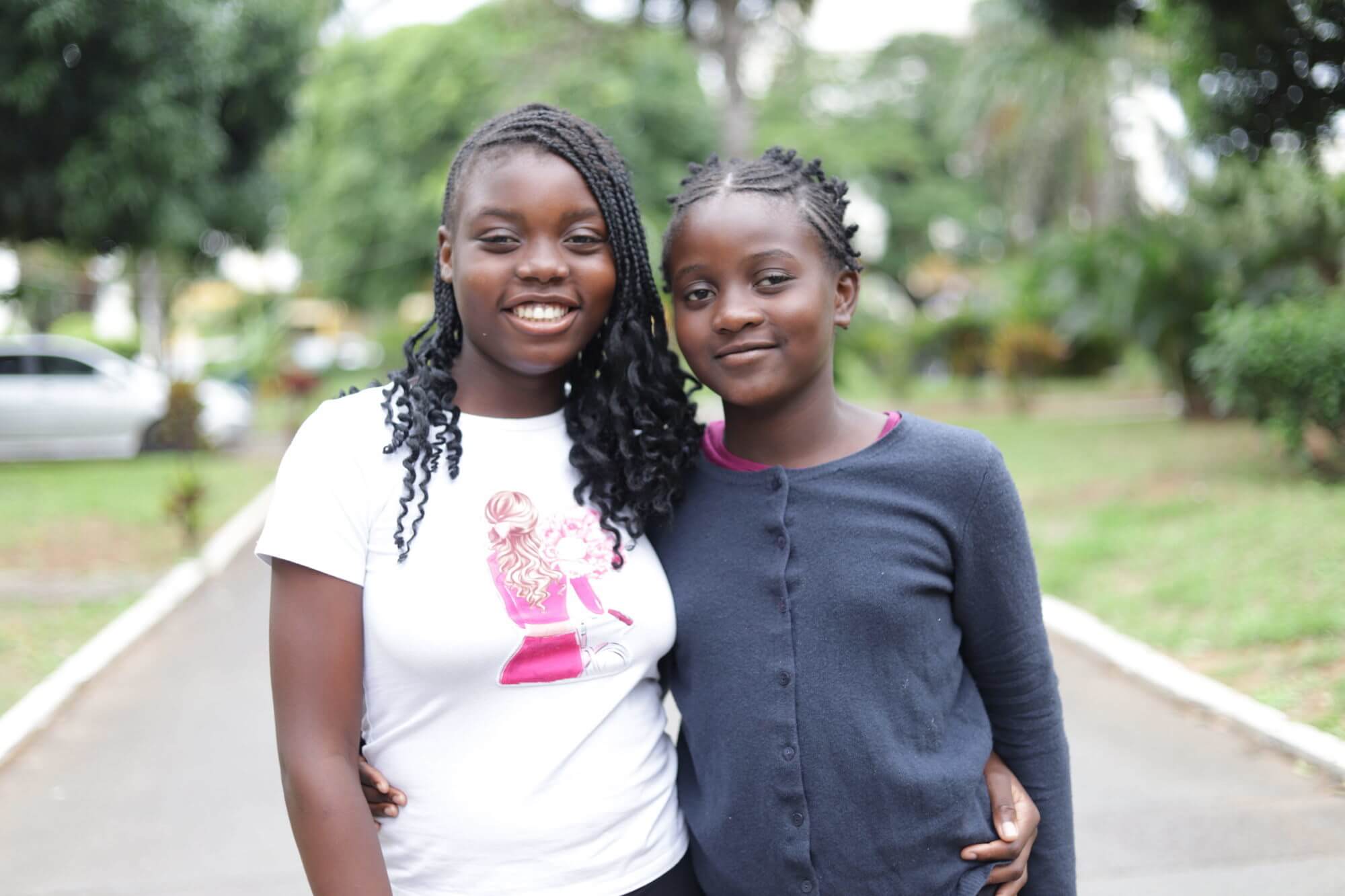 Luísa Macamo and Leona Mondlane stand together outdoors in Maputo, smiling, with Luísa's arm around Leona's shoulders, after their cataract surgeries.