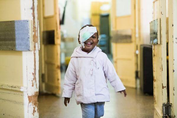 Deolinda, a small girl who received cataract surgery, smiling and looking at the camera. Her right eye is covered by a bandage.