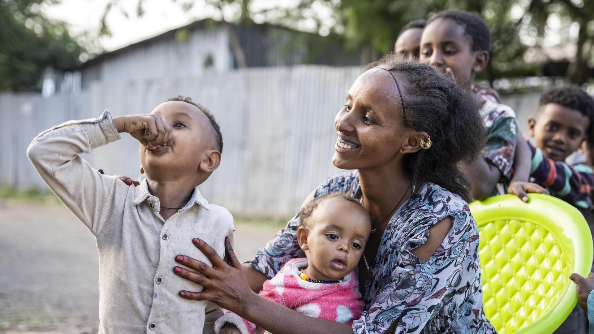 A woman sits outdoors holding a baby, with a young boy taking medicine beside her and other children gathered around, in Tigray, Ethiopia.