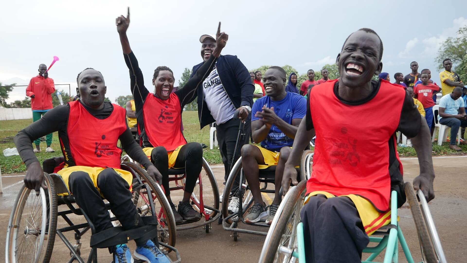 Wheelchair athletes in Ability Sports Africa bibs celebrate and cheer courtside, several with arms raised and laughing, alongside teammates.